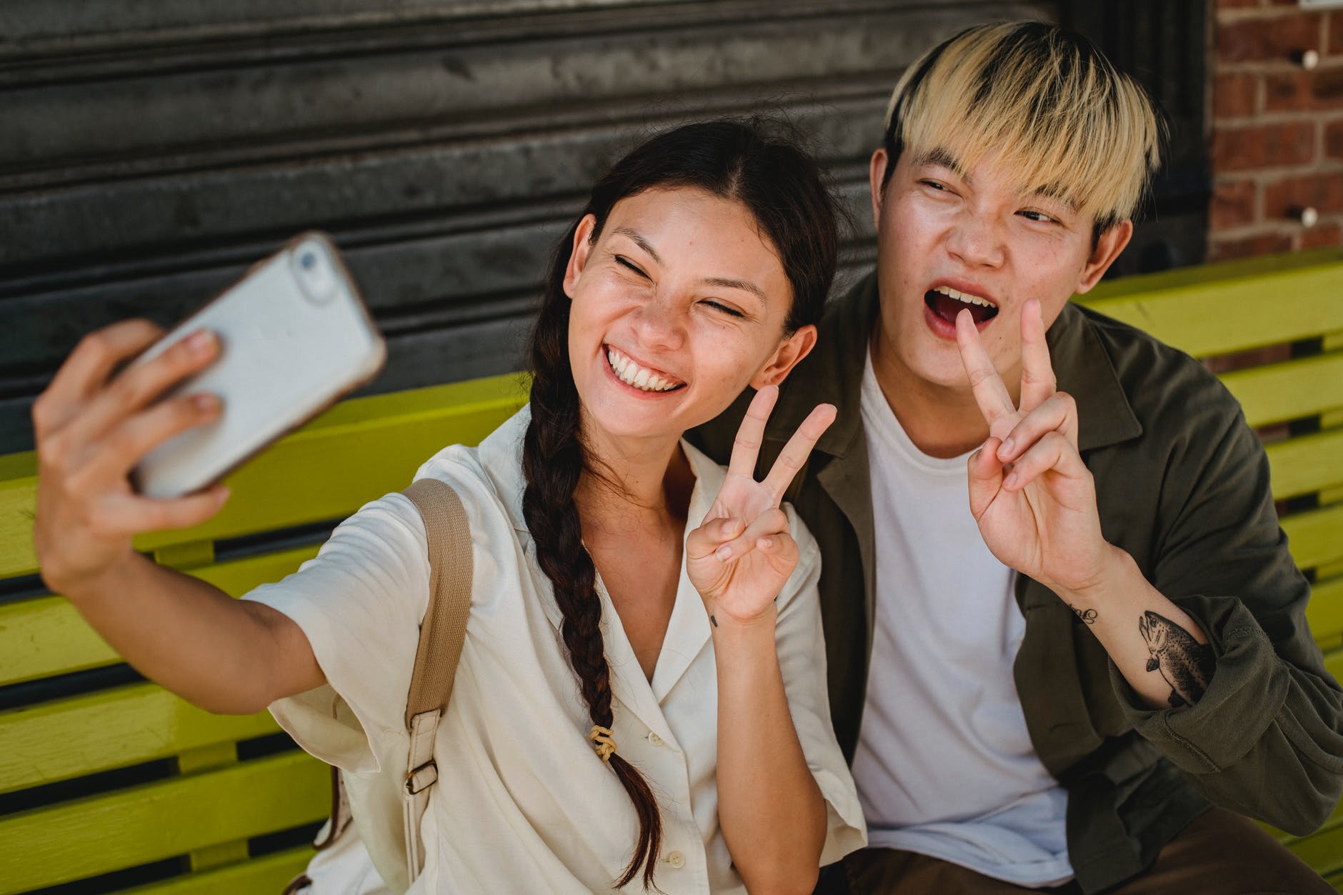 joyful asian couple taking selfies on smartphone on street bench